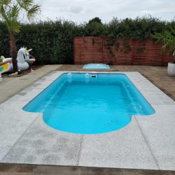 GRANITEK Marek Żoch - Niebieski basen otoczony jasnoszarą powierzchnią z kostki granitowej, w tle żywopłot, drewniany płot i dmuchany jednorożec.