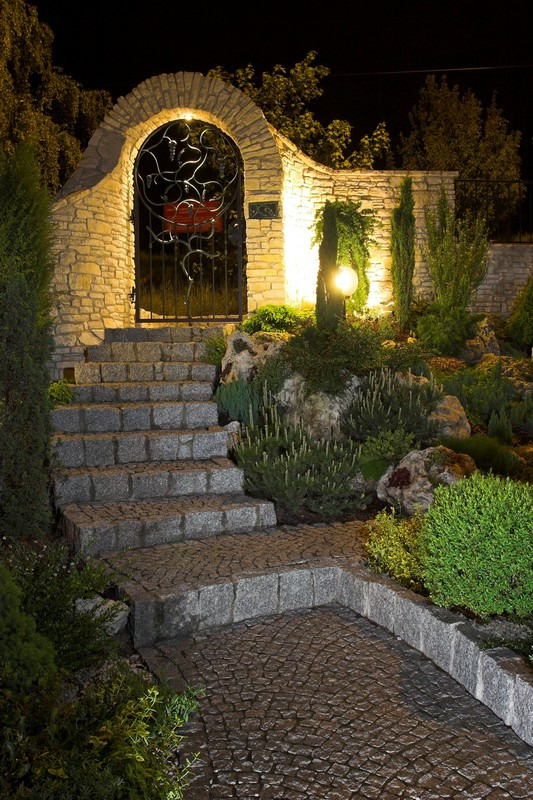 Zewnętrzne schody prowadzące do ozdobnej bramy z kamienia, oświetlone ciepłym światłem lamp, otoczone zielenią w nocnej scenerii.