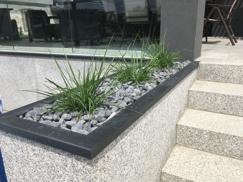 Granitowe schody zewnętrzne z donicą wypełnioną szarymi kamieniami i trawą ozdobną, widok z boku na trzy stopnie i fragment donicy.