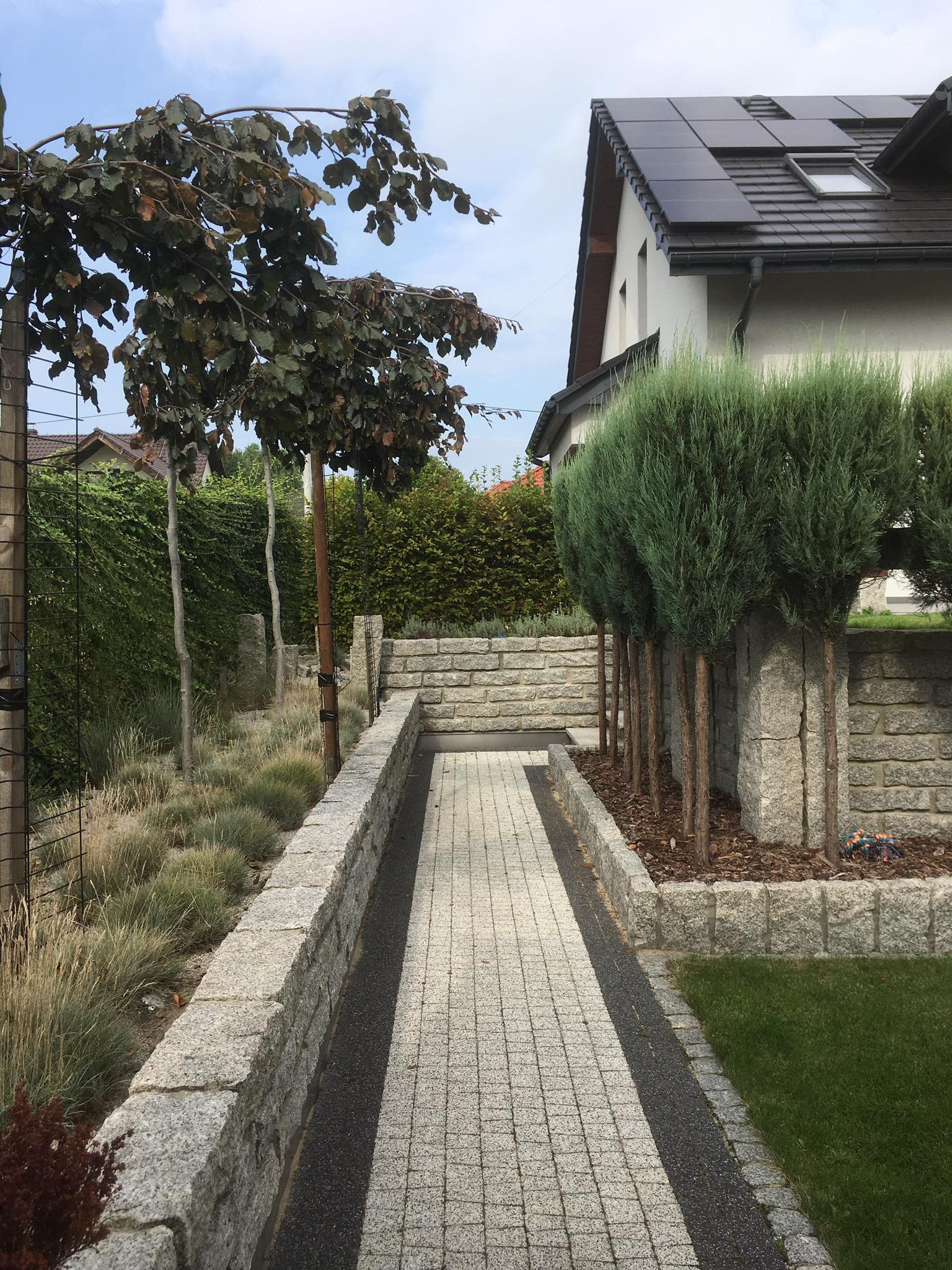 Granitowa ścieżka z jasnej kostki prowadząca przez ogród, otoczona niskimi murkami z kamienia, z drzewami po bokach i domem w tle.