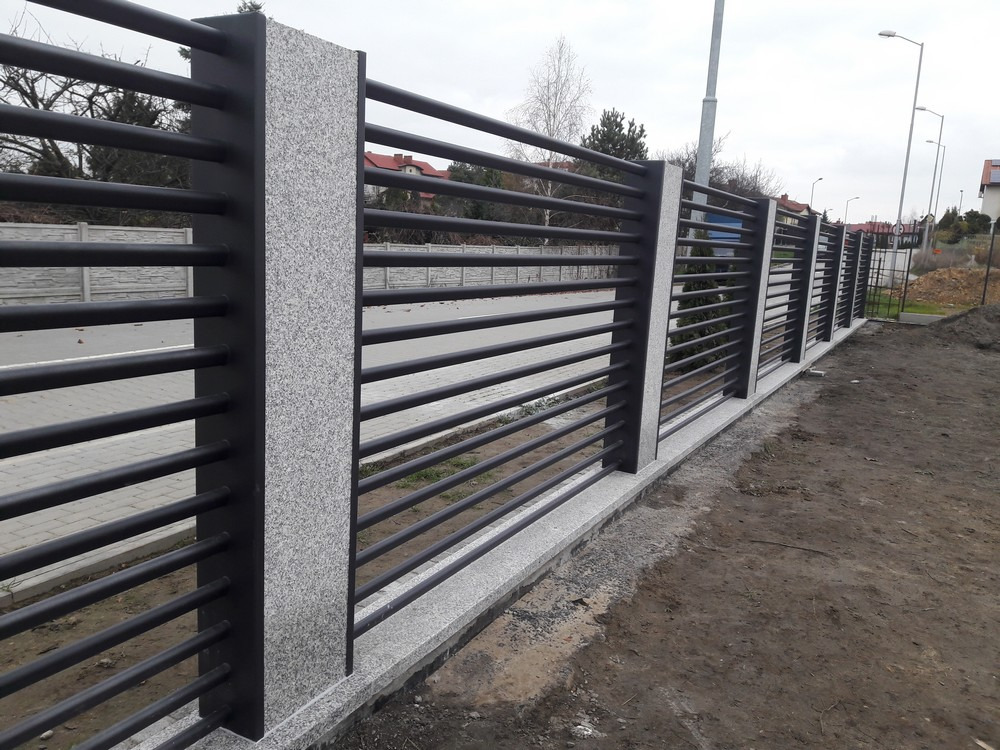 Nowoczesne ogrodzenie z poziomych, czarnych rurek stalowych i szarych, granitowych słupków, z widoczną podmurówką z tego samego materiału, na tle pochmurnego nieba i zabudowy jednorodzinnej.