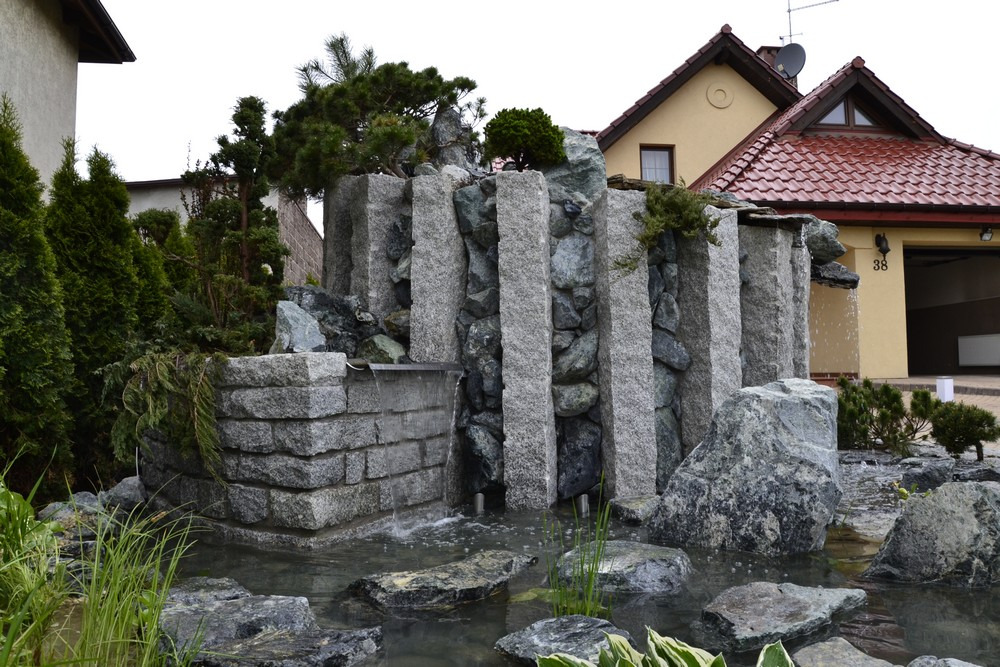 Ogród z kaskadą wodną wykonaną z pionowych, szarych bloków kamiennych i mniejszych kamieni, otoczony roślinnością i wodą, z budynkiem w tle.