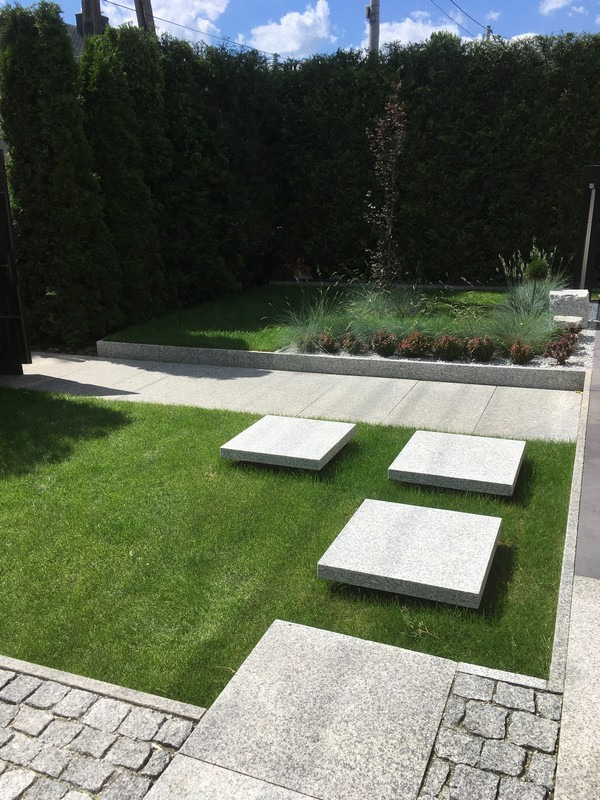 Nowoczesny ogród z kwadratowymi płytami chodnikowymi z jasnoszarego kamienia ułożonymi na trawniku, obrzeża z kostki granitowej i wysokie tuje w tle.
