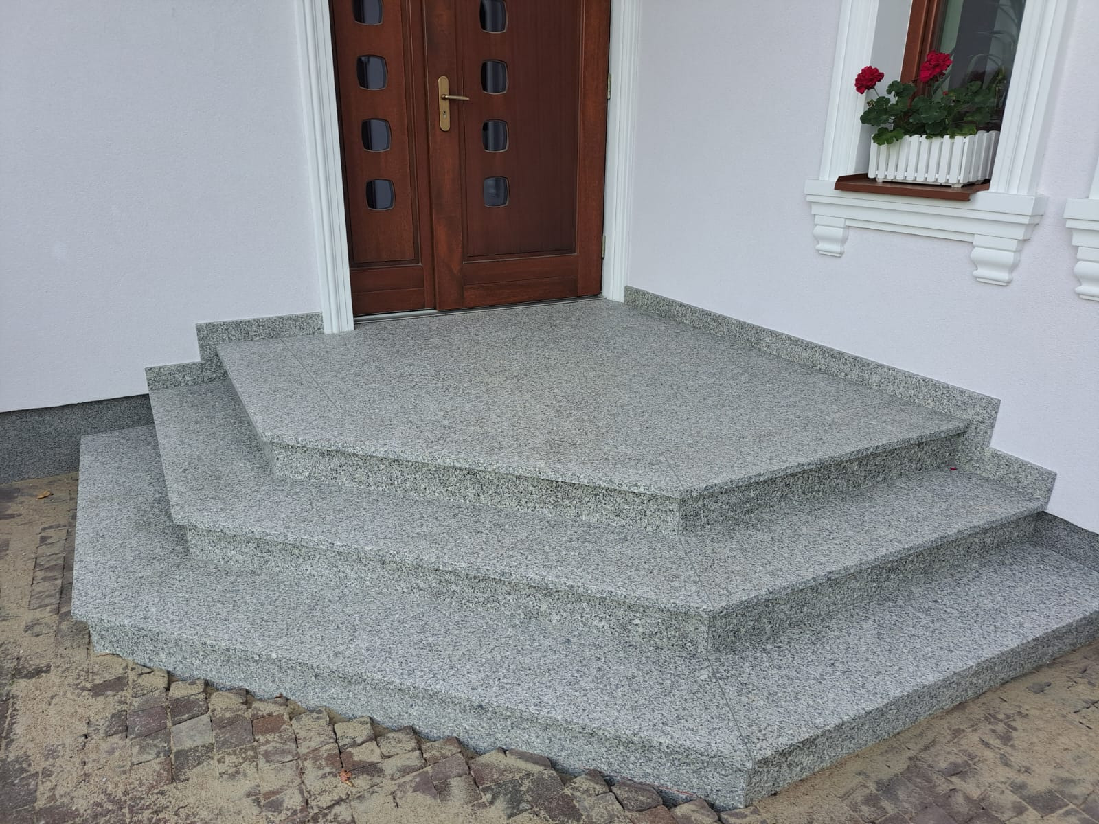 Granitowe stopnie wejściowe do domu, prowadzące do drewnianych drzwi z szybkami, z doniczką z czerwonymi kwiatami w oknie.