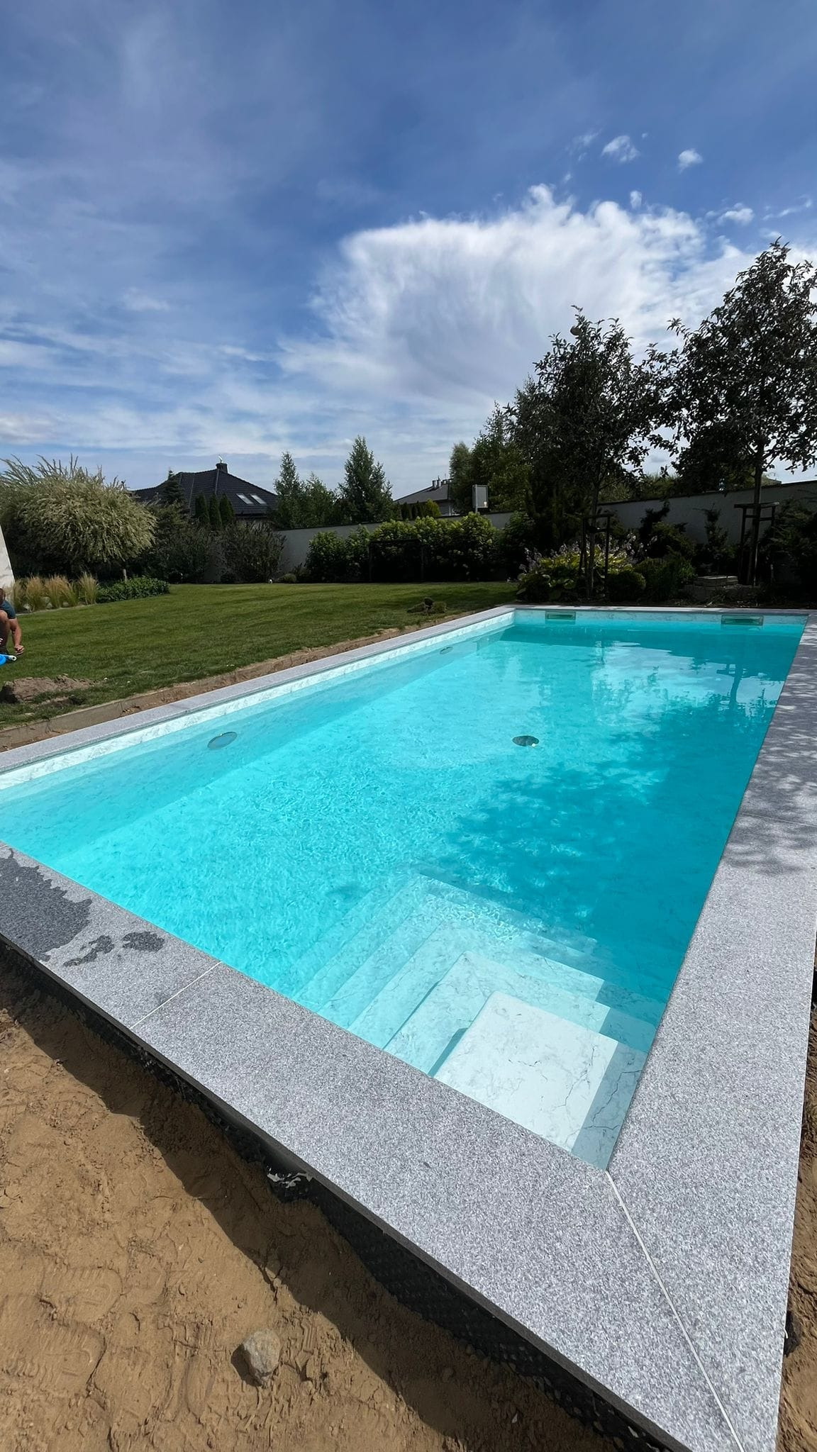 Nowoczesny basen z szarym, kamiennym obramowaniem i białymi stopniami, otoczony trawnikiem i drzewami, w słoneczny dzień.