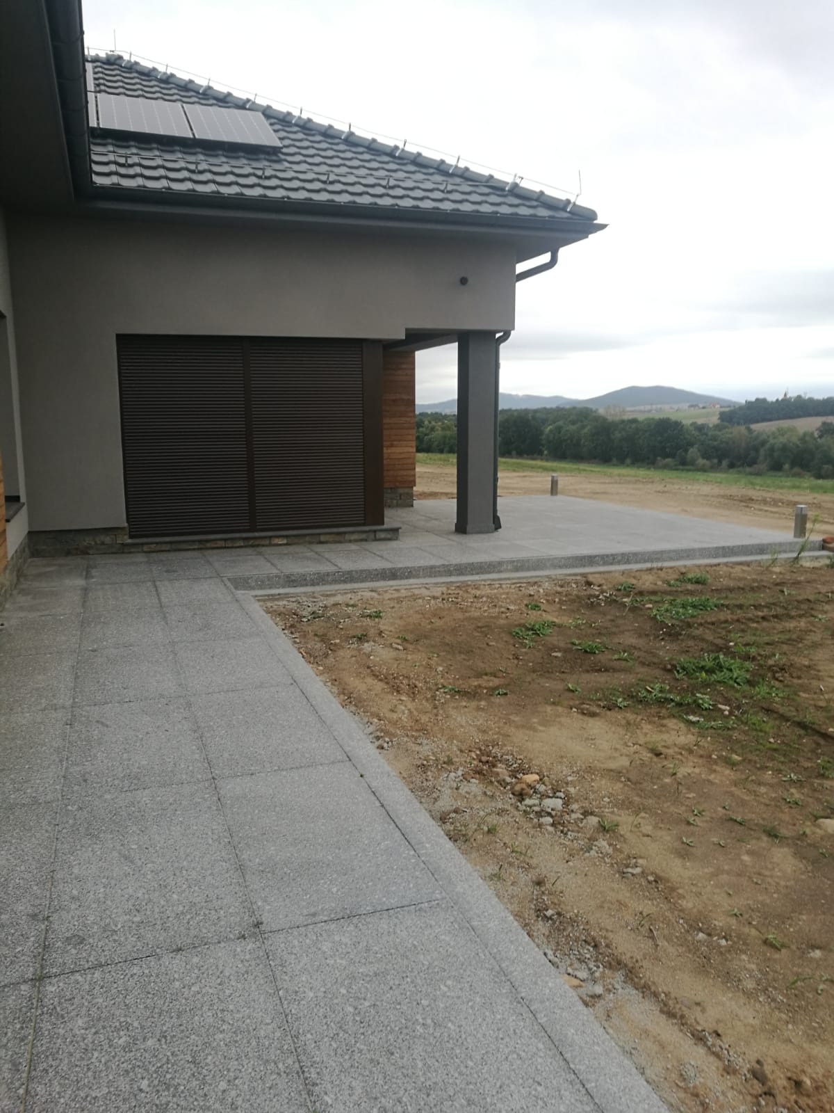 Szara, kamienna ścieżka prowadząca do tarasu z płyt granitowych, z widokiem na krajobraz pagórkowaty w tle, w pochmurny dzień.