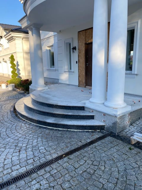 Wejście do domu z kolumnami i trzema stopniami z ciemnego kamienia, wyłożone kostką granitową w odcieniach szarości z widocznym odpływem liniowym.