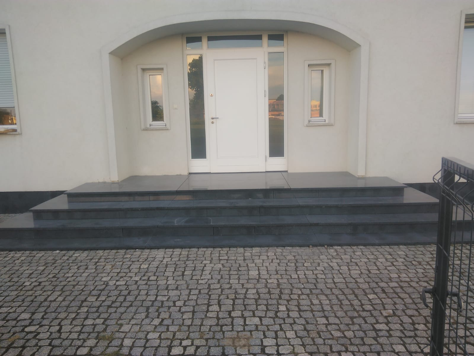 Wejście do domu z trzema stopniami wyłożonymi ciemnym kamieniem, widoczne okna boczne i bruk z kostki granitowej.
