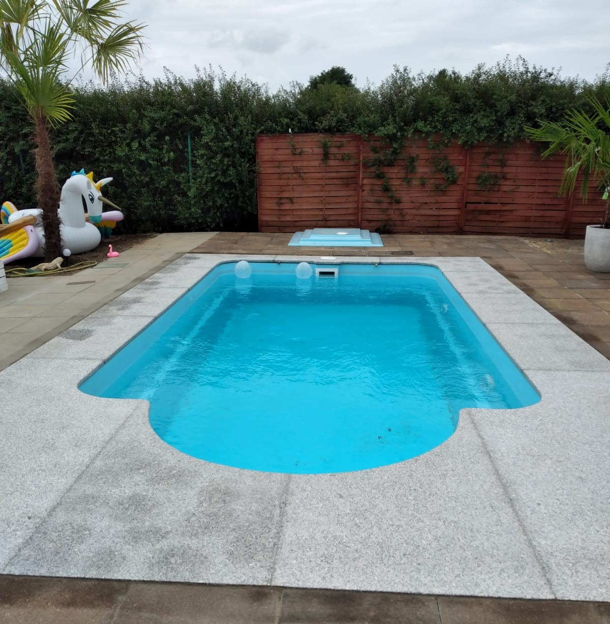 Niebieski basen otoczony jasnoszarą powierzchnią z kostki granitowej, w tle żywopłot, drewniany płot i dmuchany jednorożec.