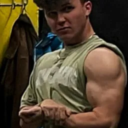 Młody mężczyzna w koszulce bez rękawów, prezentujący bicepsy w siłowni, z widocznym w tle wieszakiem na ubrania i żółtą ścianą.