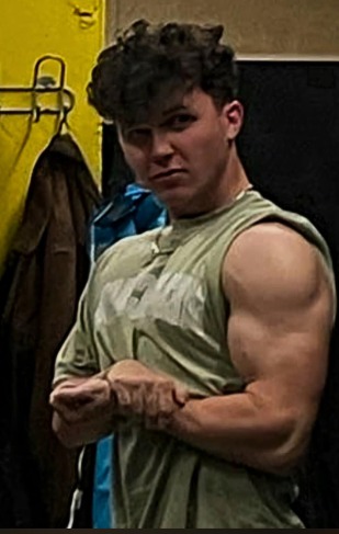 Młody mężczyzna w koszulce bez rękawów, prezentujący bicepsy w siłowni, z widocznym w tle wieszakiem na ubrania i żółtą ścianą.