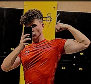 Młody mężczyzna robi selfie w lustrze, prezentując bicepsy w pomarańczowej koszulce na tle szafek w szatni.