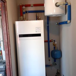 Krzysztof Solecki - Biała pompa ciepła Panasonic z zbiornikiem buforowym i niebieskimi rurami w ciasnym pomieszczeniu technicznym, widoczne również czerwone rury, manometry i instalacja w ścianie