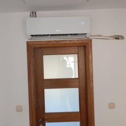 Krzysztof Solecki - Biały klimatyzator marki Gree zamontowany nad drewnianymi drzwiami z trzema szybami, widoczne wystające rury instalacyjne i wyświetlacz wskazujący temperaturę 22 stopnie.