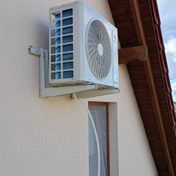 Krzysztof Solecki - Zewnętrzna jednostka klimatyzatora zamontowana na elewacji domu, widoczny przewód odprowadzający skropliny i fragment okna zabezpieczonego folią.