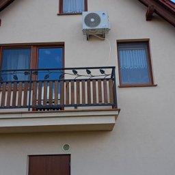 Krzysztof Solecki - Biała jednostka zewnętrzna klimatyzacji zamontowana na elewacji domu z balkonem i brązowymi oknami, widoczna instalacja elektryczna.