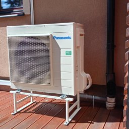 Krzysztof Solecki - Zewnętrzna jednostka klimatyzacji Panasonic na metalowym stelażu, zainstalowana na drewnianym tarasie przy ścianie budynku.