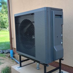 Krzysztof Solecki - Szara pompa ciepła powietrze-woda zamontowana na czarnej, metalowej podstawie na betonowym fundamencie przy ścianie budynku.