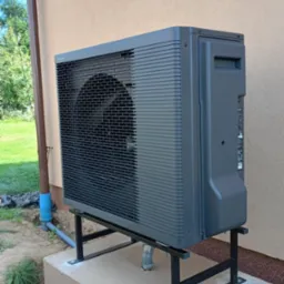 Szara pompa ciepła powietrze-woda zamontowana na czarnej, metalowej podstawie na betonowym fundamencie przy ścianie budynku.