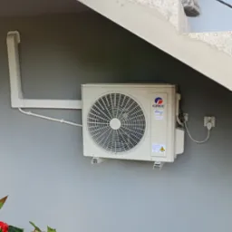 Zamontowana jednostka zewnętrzna klimatyzacji Gree z białymi rurami, widoczna spod schodów, z czerwonymi kwiatami w lewym dolnym rogu.