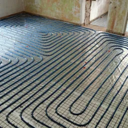 Niebieskie rury ogrzewania podłogowego ułożone na siatce w surowym wnętrzu. Widoczne przyłącza i fragment okna. Realizacja instalacji ogrzewania podłogowego.