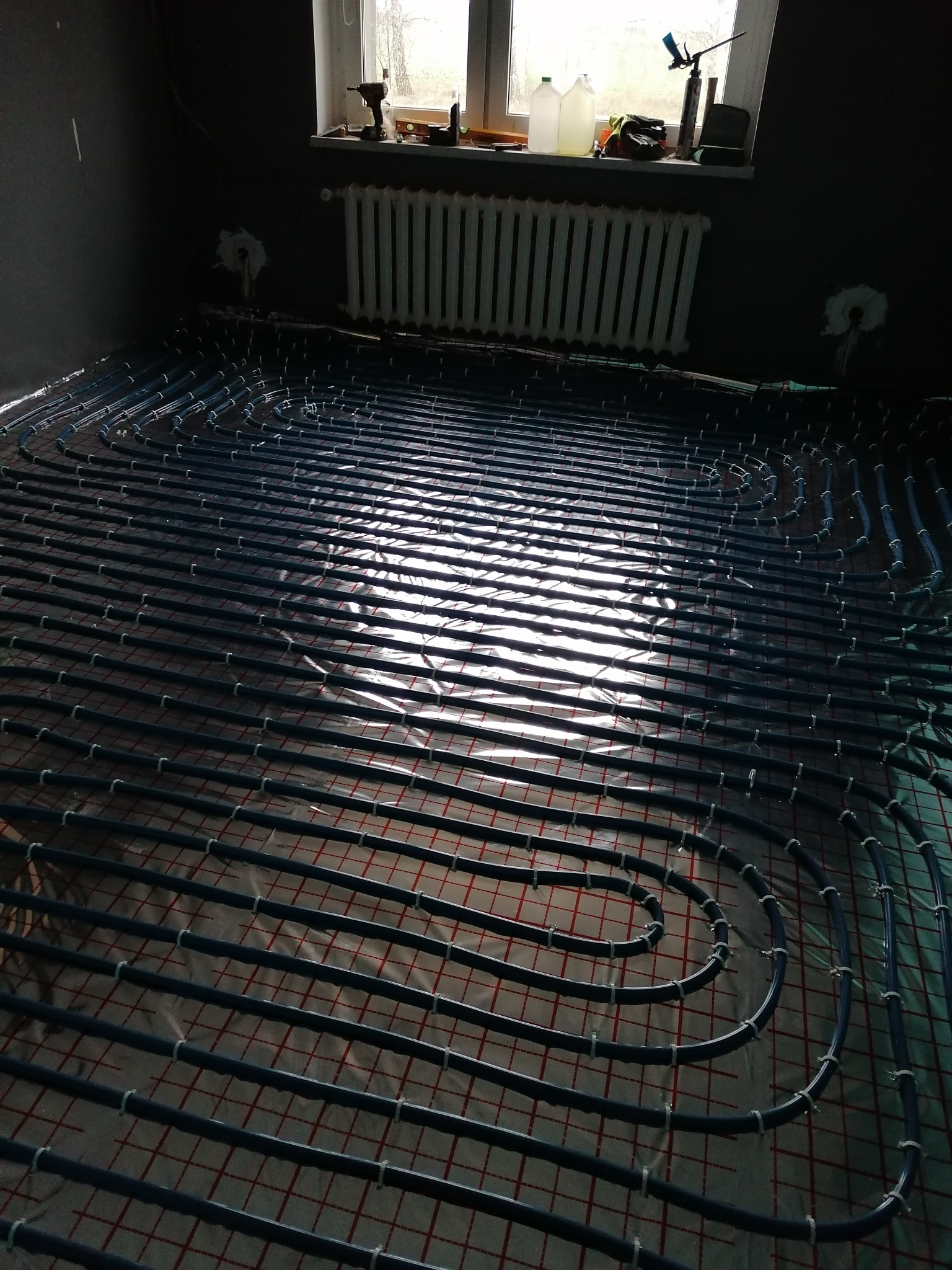 Rozłożone rury systemu ogrzewania podłogowego na folii z siatką w nowym pomieszczeniu z szarą ścianą i grzejnikiem pod oknem, widoczne narzędzia na parapecie.