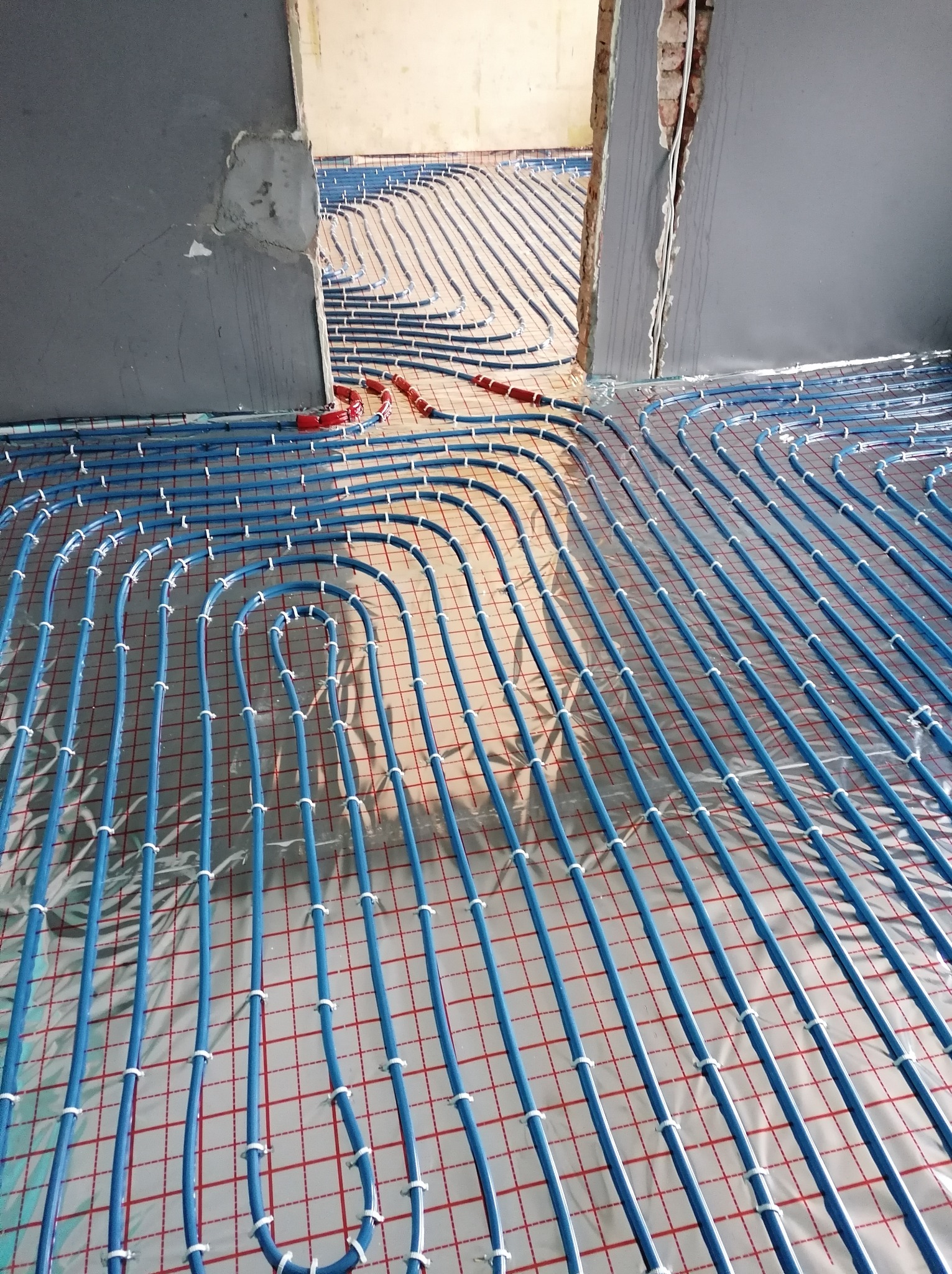 Niebieskie rury systemu grzewczego ułożone na folii z czerwoną kratą, w surowym stanie, widoczne fragmenty ścian w trakcie prac wykończeniowych.