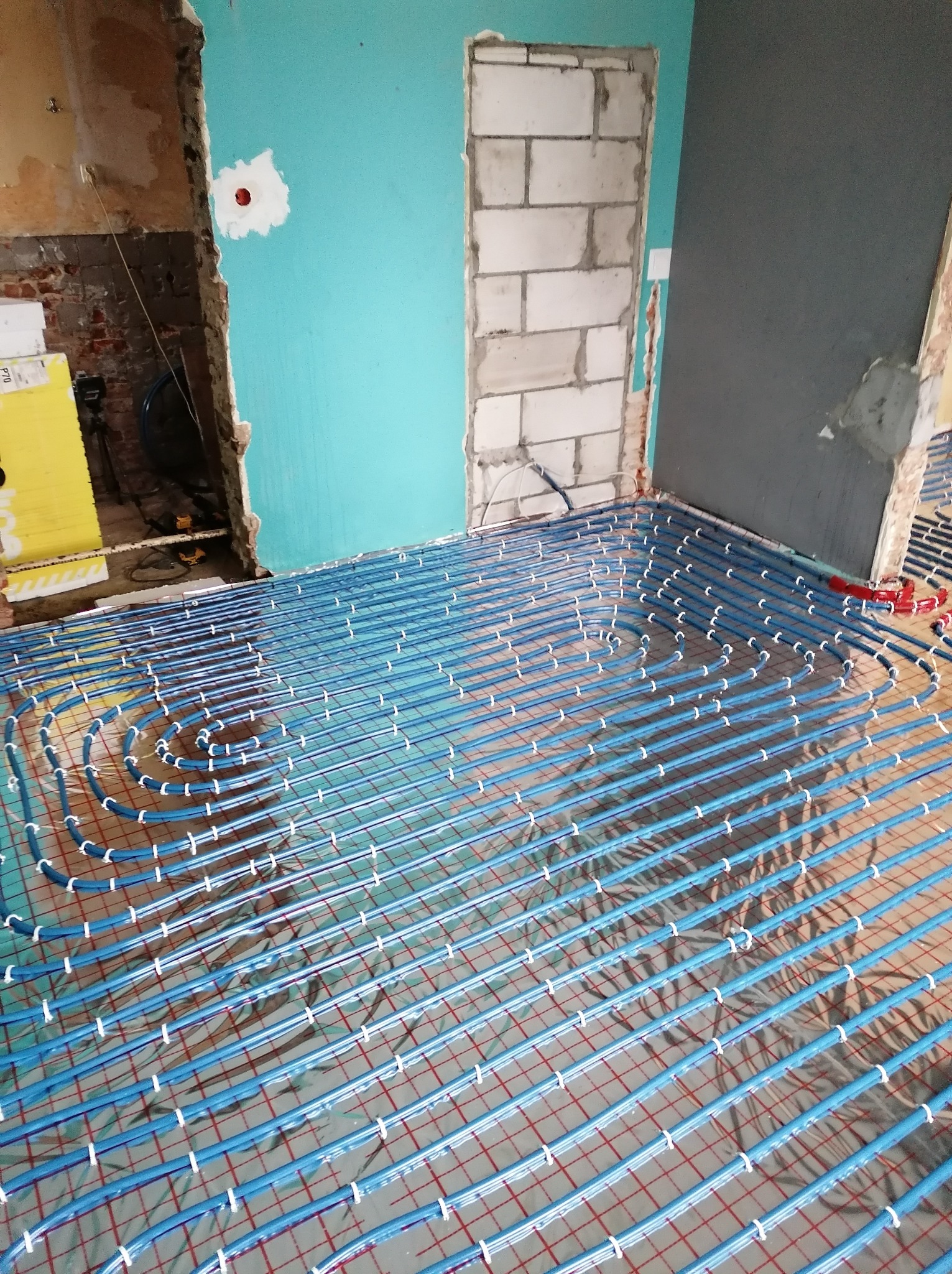 Niebieskie rurki instalacji grzewczej układane spiralnie na folii odblaskowej w surowym wnętrzu z fragmentami ścian w kolorze turkusowym i szarym, widoczne niedokończone ściany z cegieł i bloczków.