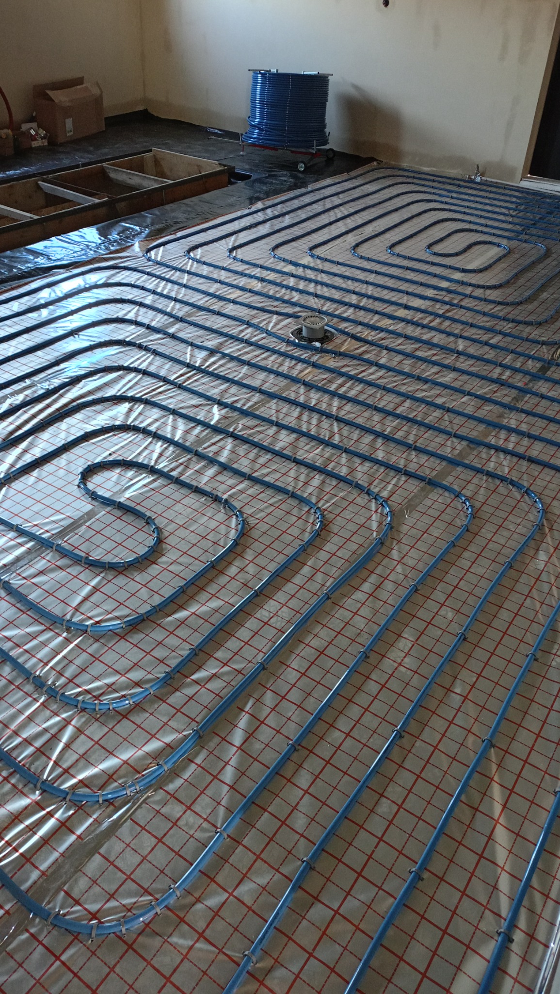 Niebieskie rurki systemu grzewczego ułożone spiralnie na folii z czerwoną kratką, widoczny kolektor podłogowy i rolka rur na wózku w tle.