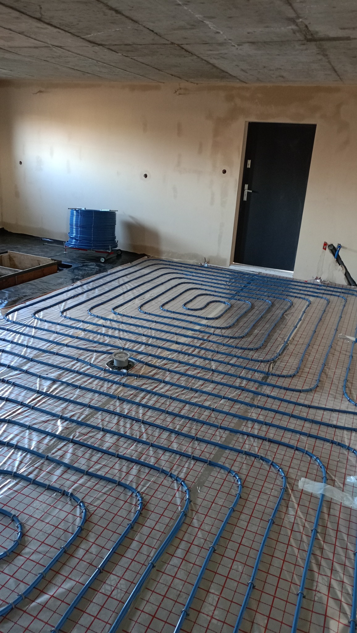 Niebieskie rury ułożone spiralnie na podłodze z siatką, stanowiące instalację ogrzewania podłogowego w surowym pomieszczeniu z betonowym sufitem i przygotowaną ścianą.