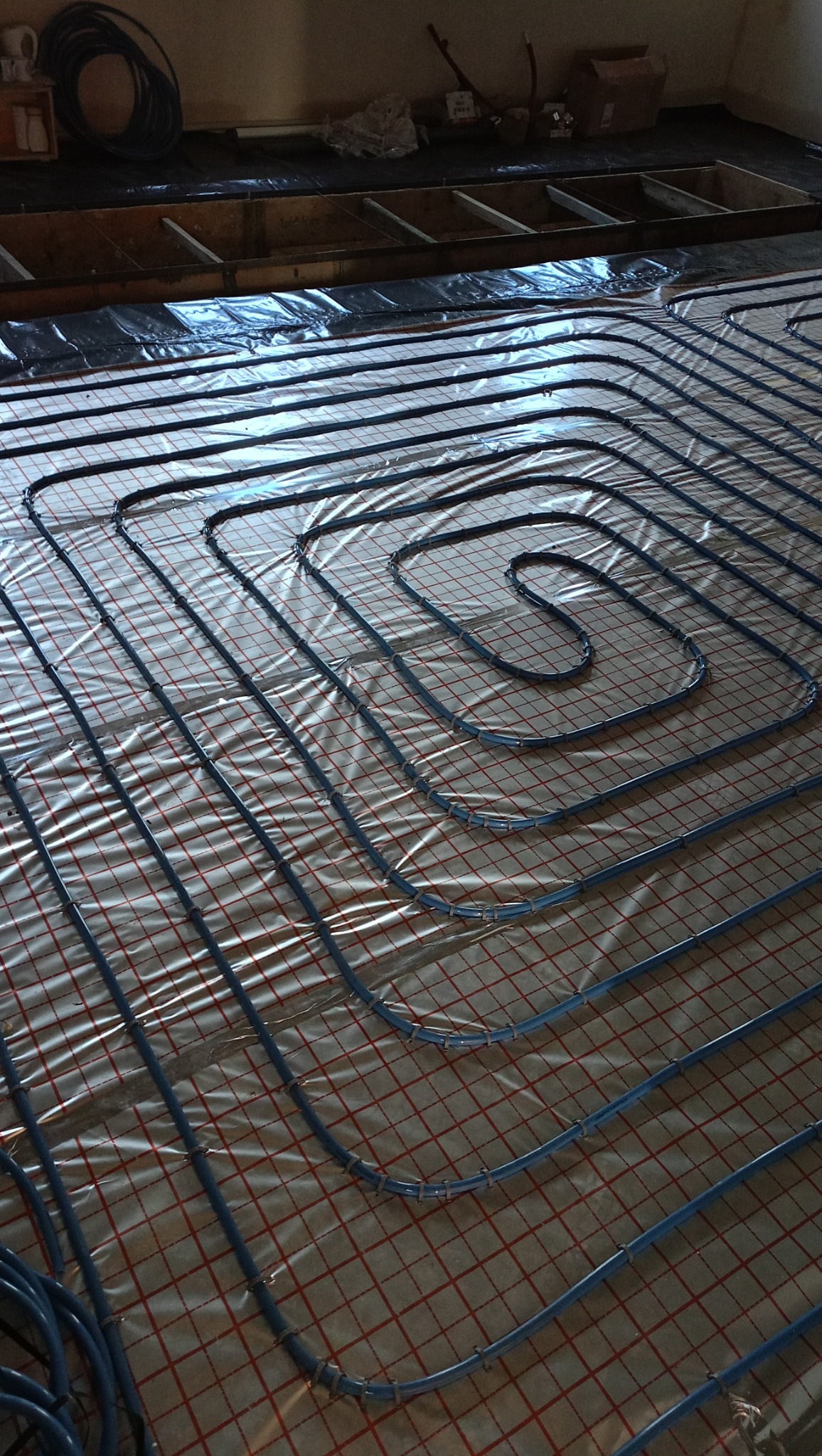 Układanie niebieskich rur systemu grzewczego na folii z czerwoną siatką na podłodze w trakcie instalacji.