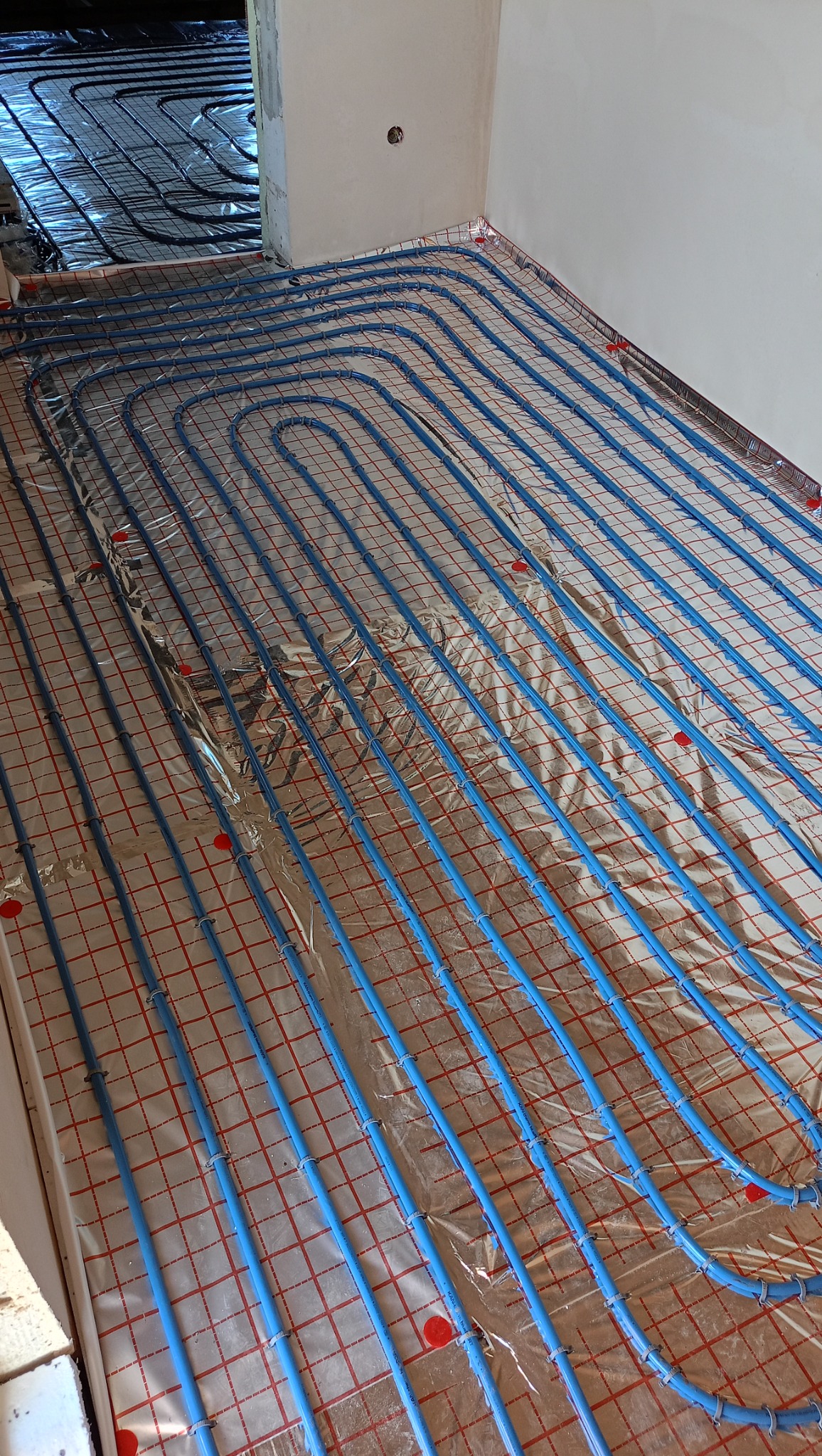 Niebieskie rury ogrzewania podłogowego ułożone na folii izolacyjnej z czerwoną siatką, widok z góry na etapie instalacji, fragment ściany w tle.