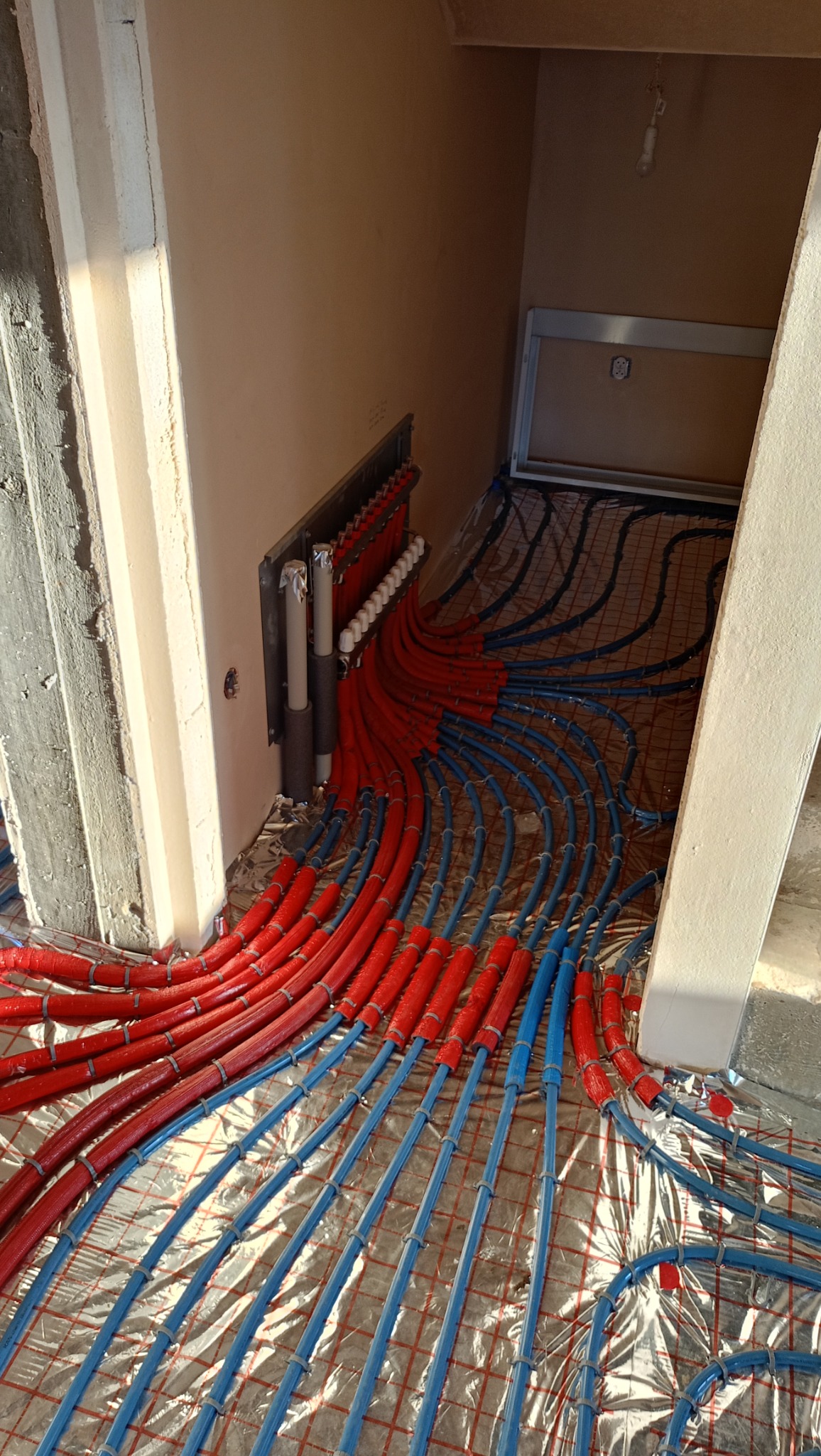 Instalacja ogrzewania podłogowego z czerwonymi i niebieskimi rurkami ułożonymi na folii izolacyjnej i siatce w pomieszczeniu w trakcie prac wykończeniowych.