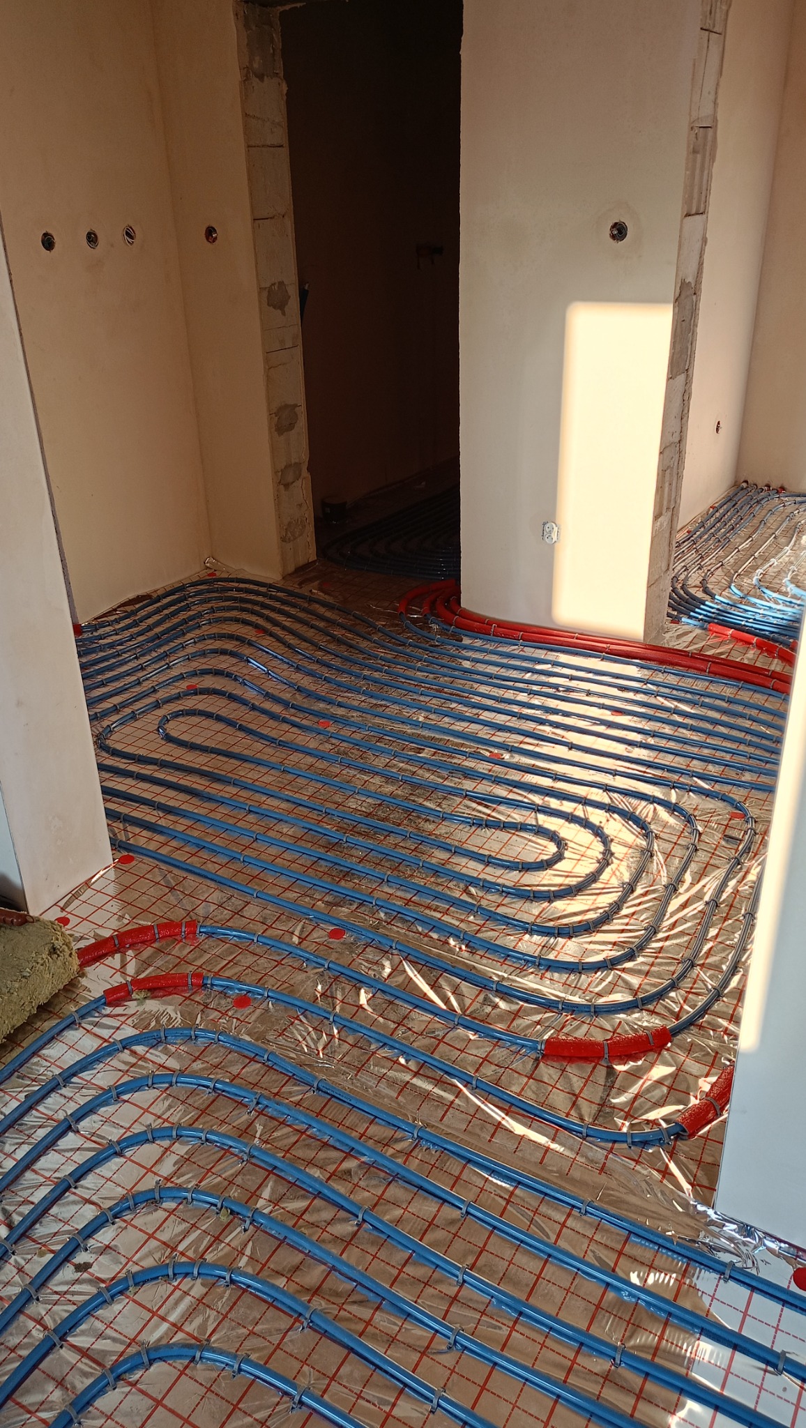 Niebieskie rury systemu ogrzewania podłogowego ułożone na folii izolacyjnej w surowym wnętrzu, widoczne fragmenty ścian i otwór drzwiowy w stanie deweloperskim.