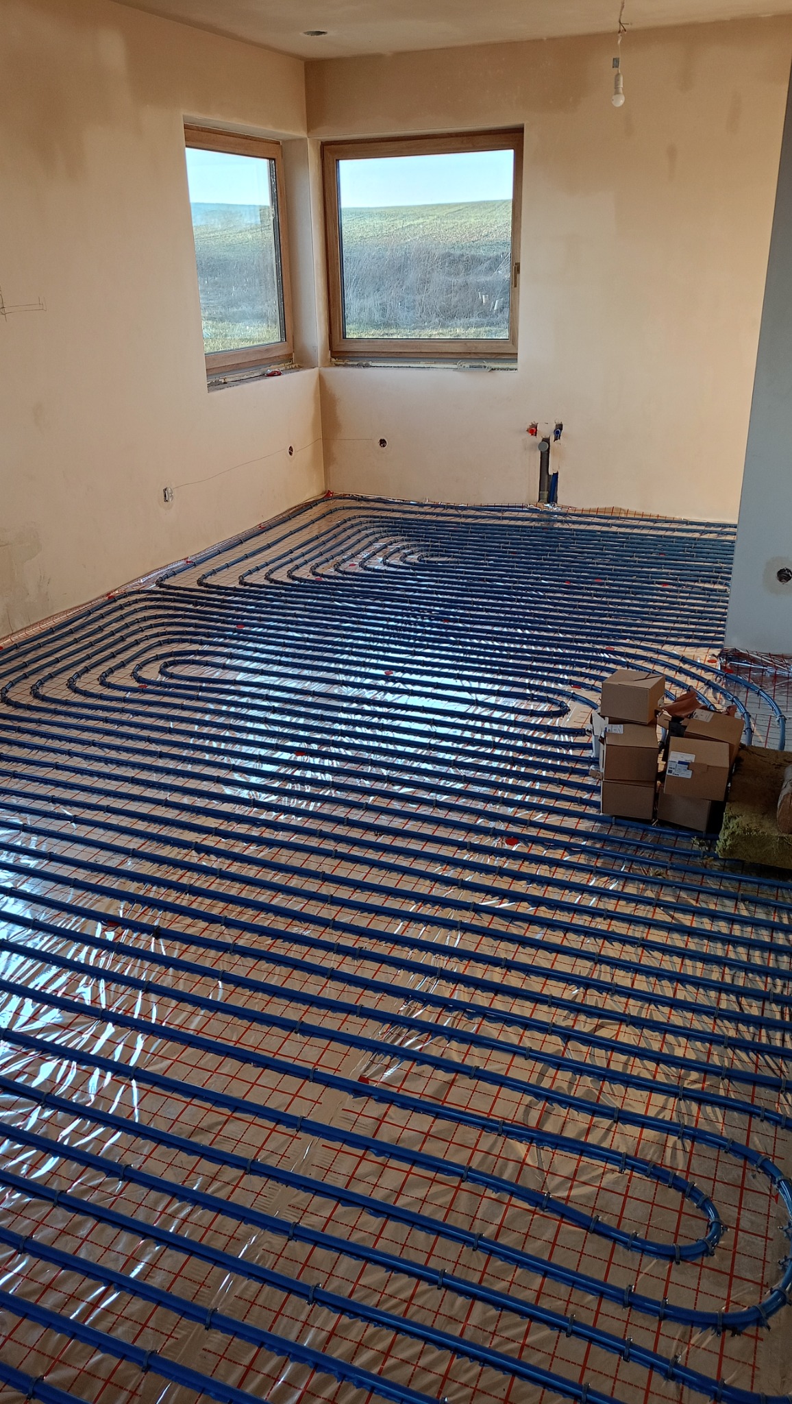 Niebieskie rurki ogrzewania podłogowego ułożone na folii w surowym wnętrzu z dwoma oknami, widoczne kartony i wystające rury instalacji.