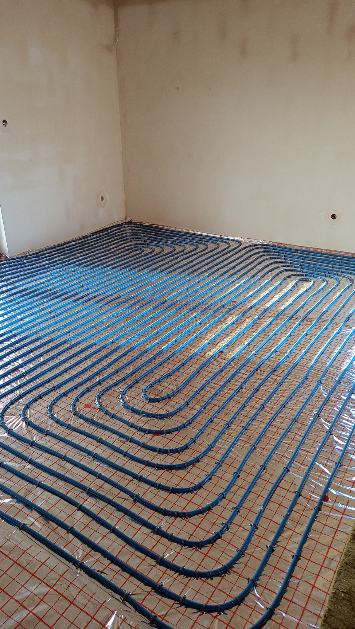 Niebieskie rury ogrzewania podłogowego ułożone w pętle na folii z czerwoną siatką w surowym wnętrzu przed wylewką.