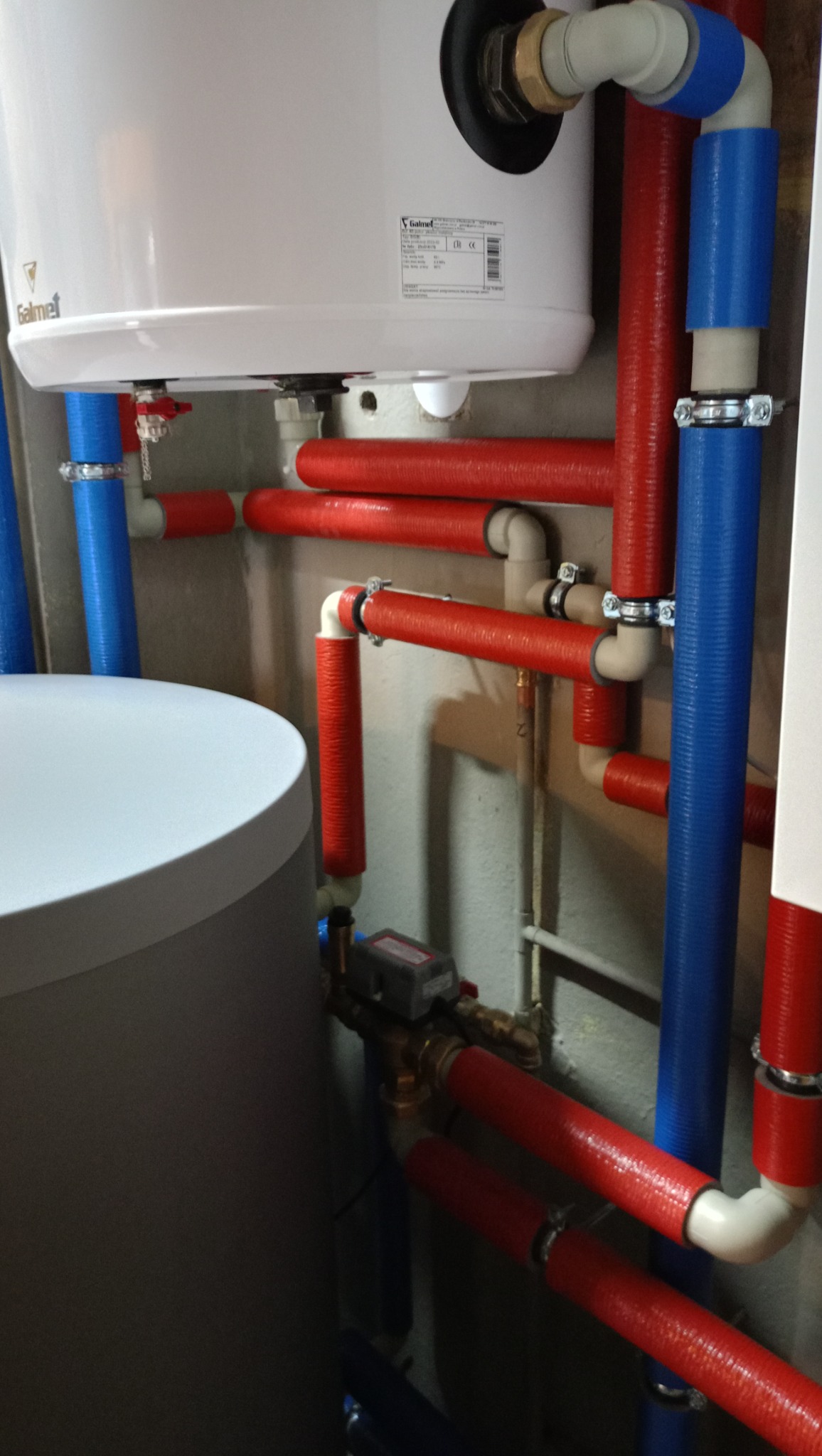 Węzeł instalacyjny z białym zasobnikiem i rurami w izolacji termicznej w kolorze czerwonym i niebieskim, zamontowany przy szarej ścianie.