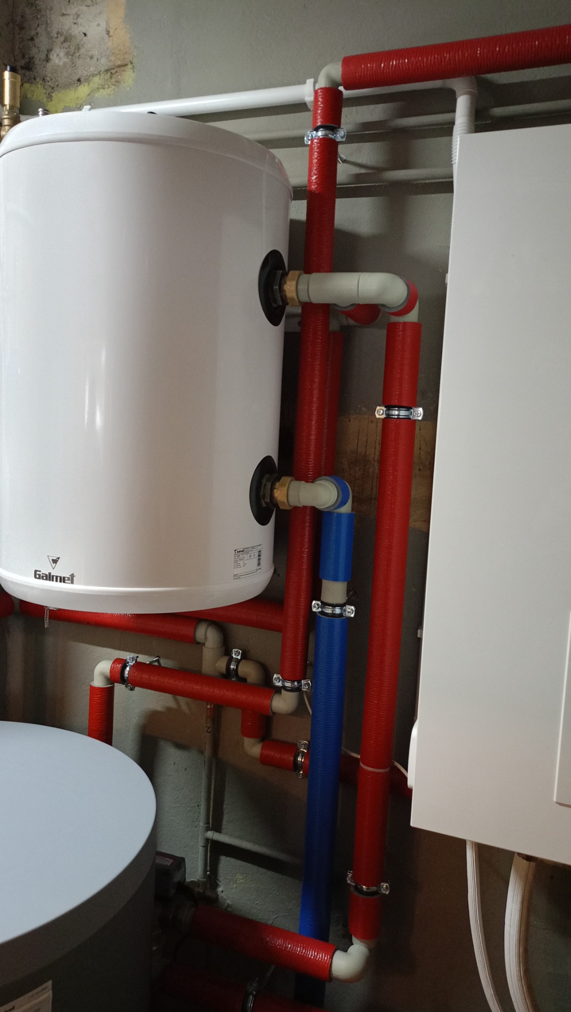 Biały zbiornik Galmet z podłączeniami rur w izolacji termicznej, czerwonej i niebieskiej, widoczne na tle szarej ściany w kotłowni.