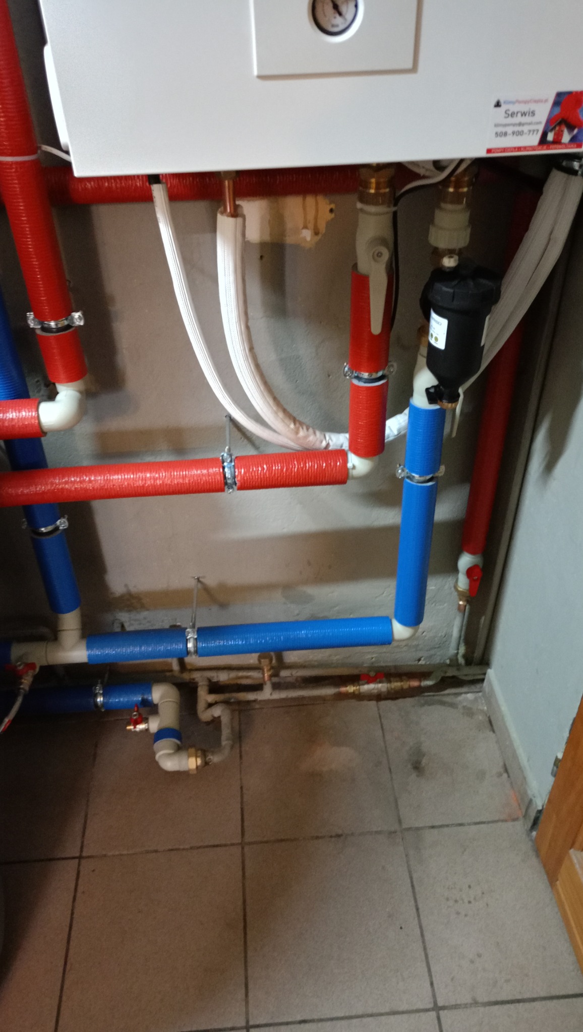 Instalacja grzewcza z rurami w izolacji czerwonej i niebieskiej, zaworami i filtrem, zamontowana przy ścianie i podłączona do białego urządzenia z manometrem.