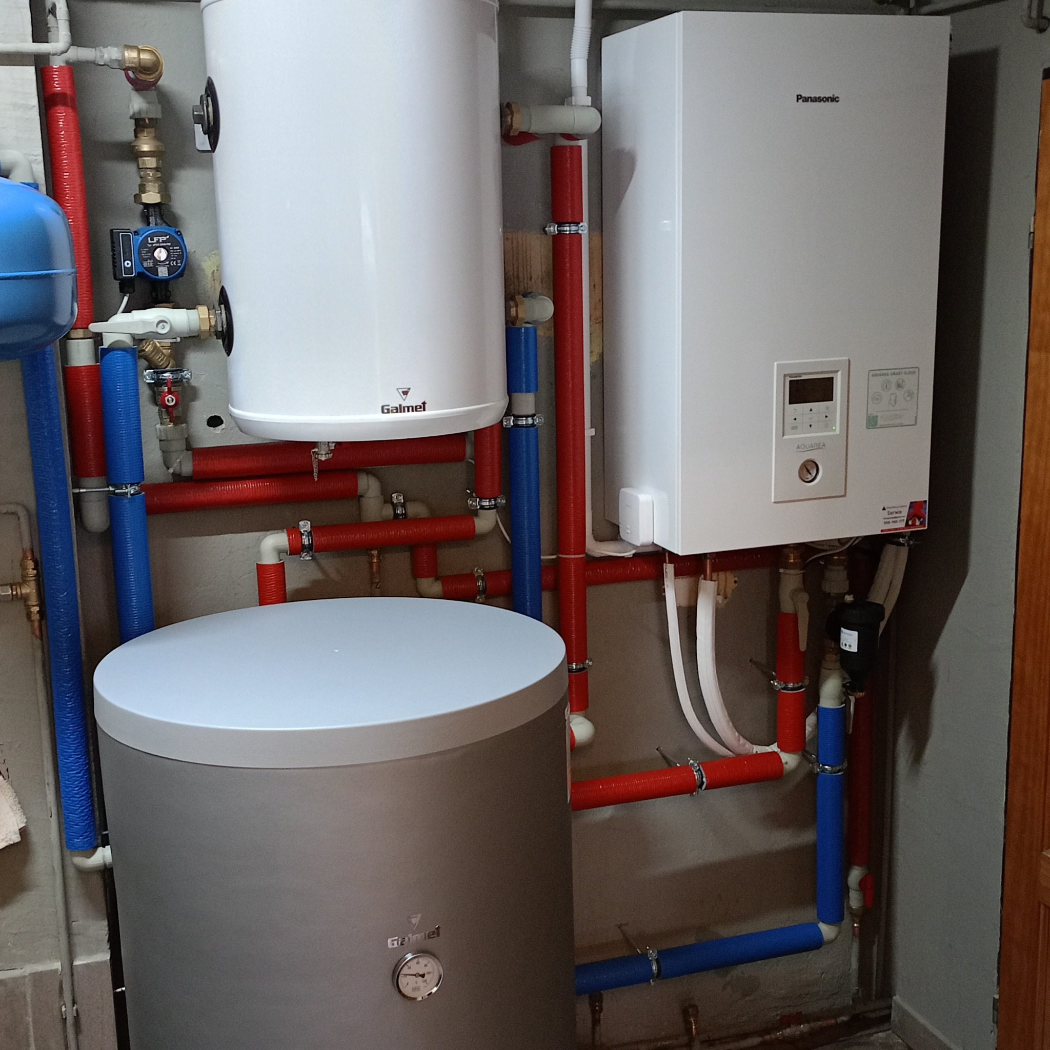 Instalacja grzewcza w piwnicy: biały zasobnik Galmet, szary zbiornik i biała jednostka Panasonic Aquarea z rurami w izolacji czerwonej i niebieskiej.