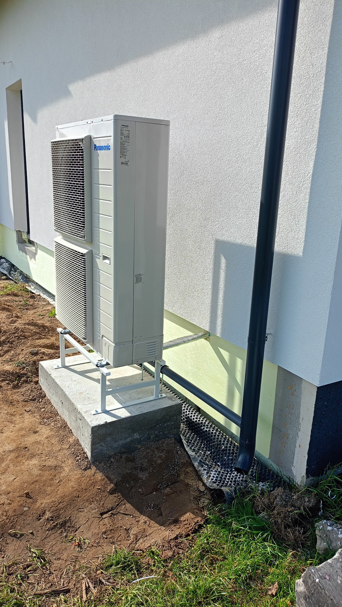 Biała pompa ciepła Panasonic na betonowej podstawie, zamontowana przy ścianie budynku. Widoczna rynna odprowadzająca wodę. Trawa i ziemia wokół.