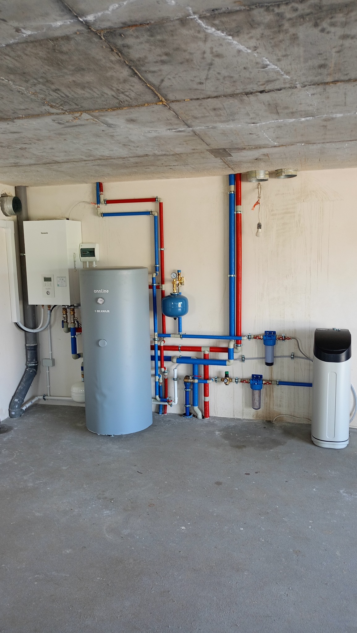 Kompleksowa instalacja grzewcza z szarym zbiornikiem Biwar, filtrami i rurami w kolorach czerwonym i niebieskim na tle surowego sufitu.