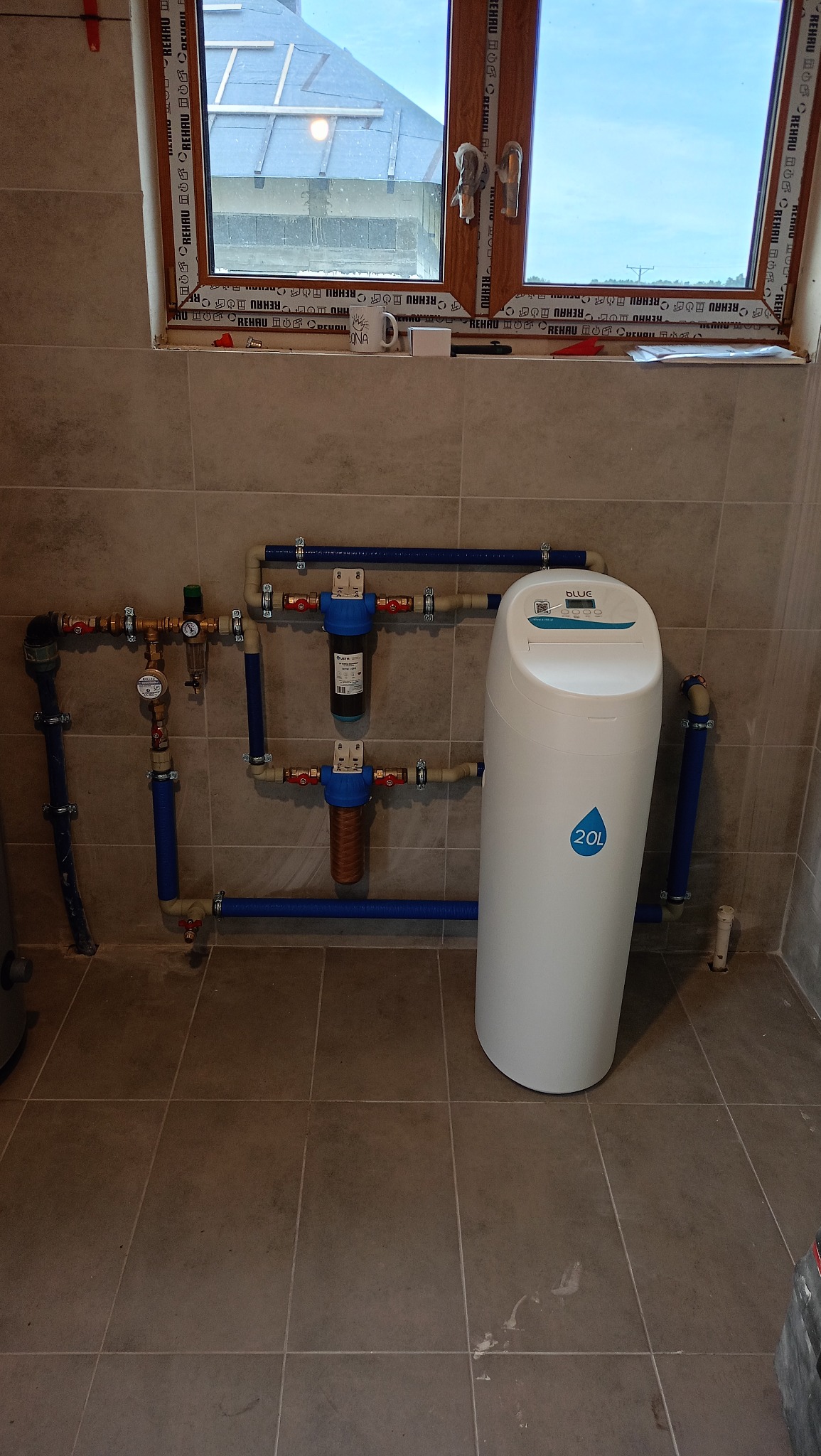Instalacja filtrów wody z niebieskimi rurami i białym zmiękczaczem wody 20L Blue, zamontowana na kafelkowanej ścianie pod oknem, widok z przodu.