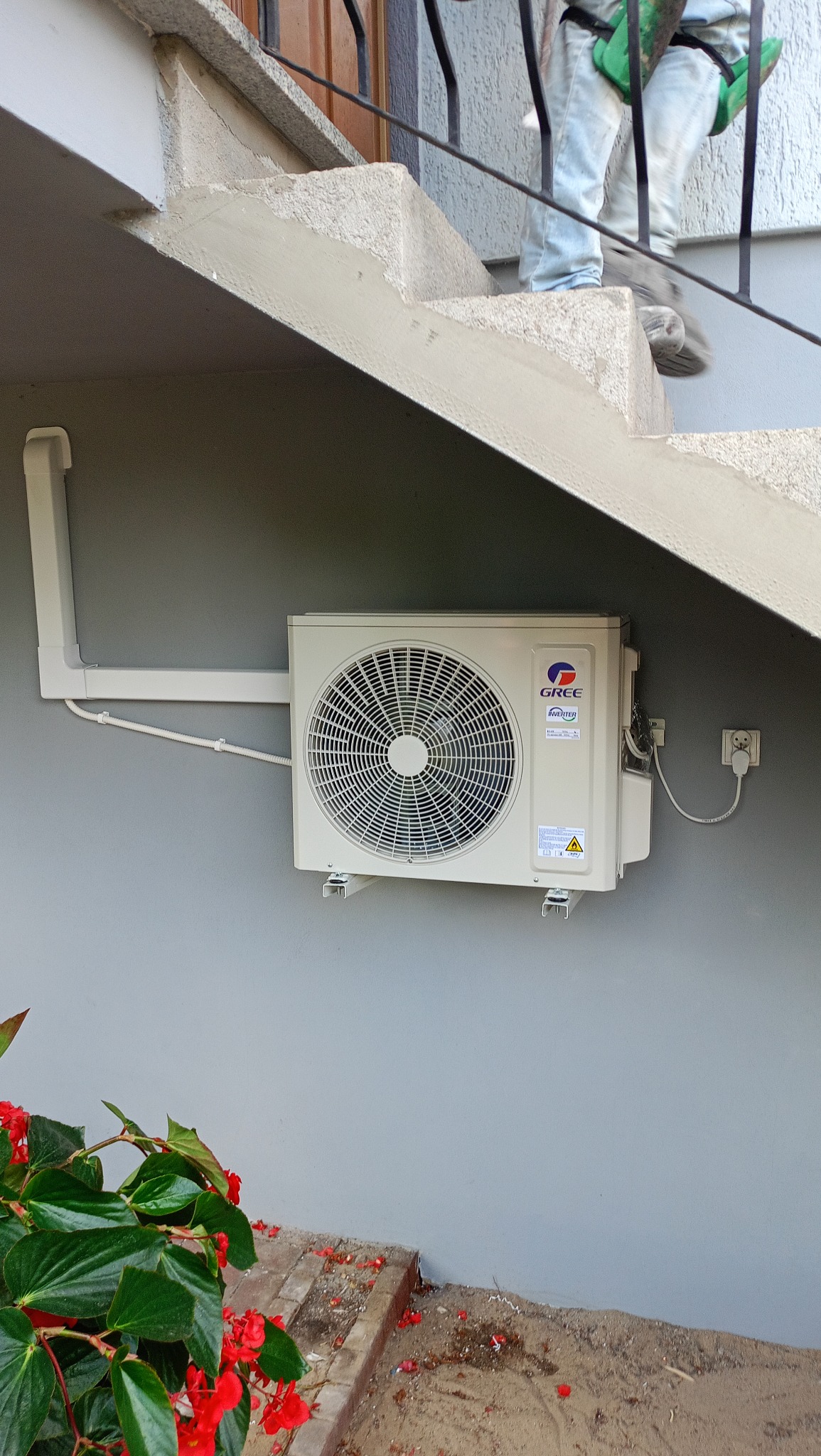 Zamontowana jednostka zewnętrzna klimatyzacji Gree z białymi rurami, widoczna spod schodów, z czerwonymi kwiatami w lewym dolnym rogu.