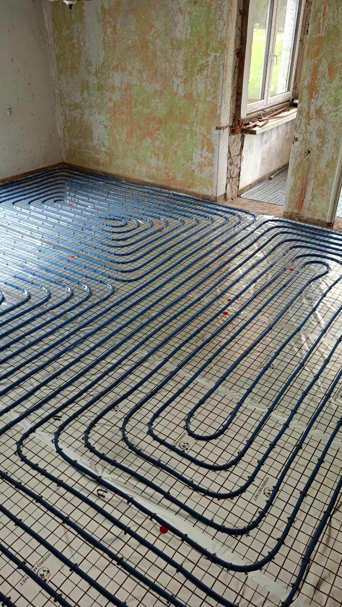 Niebieskie rury ogrzewania podłogowego ułożone na siatce w surowym wnętrzu. Widoczne przyłącza i fragment okna. Realizacja instalacji ogrzewania podłogowego.