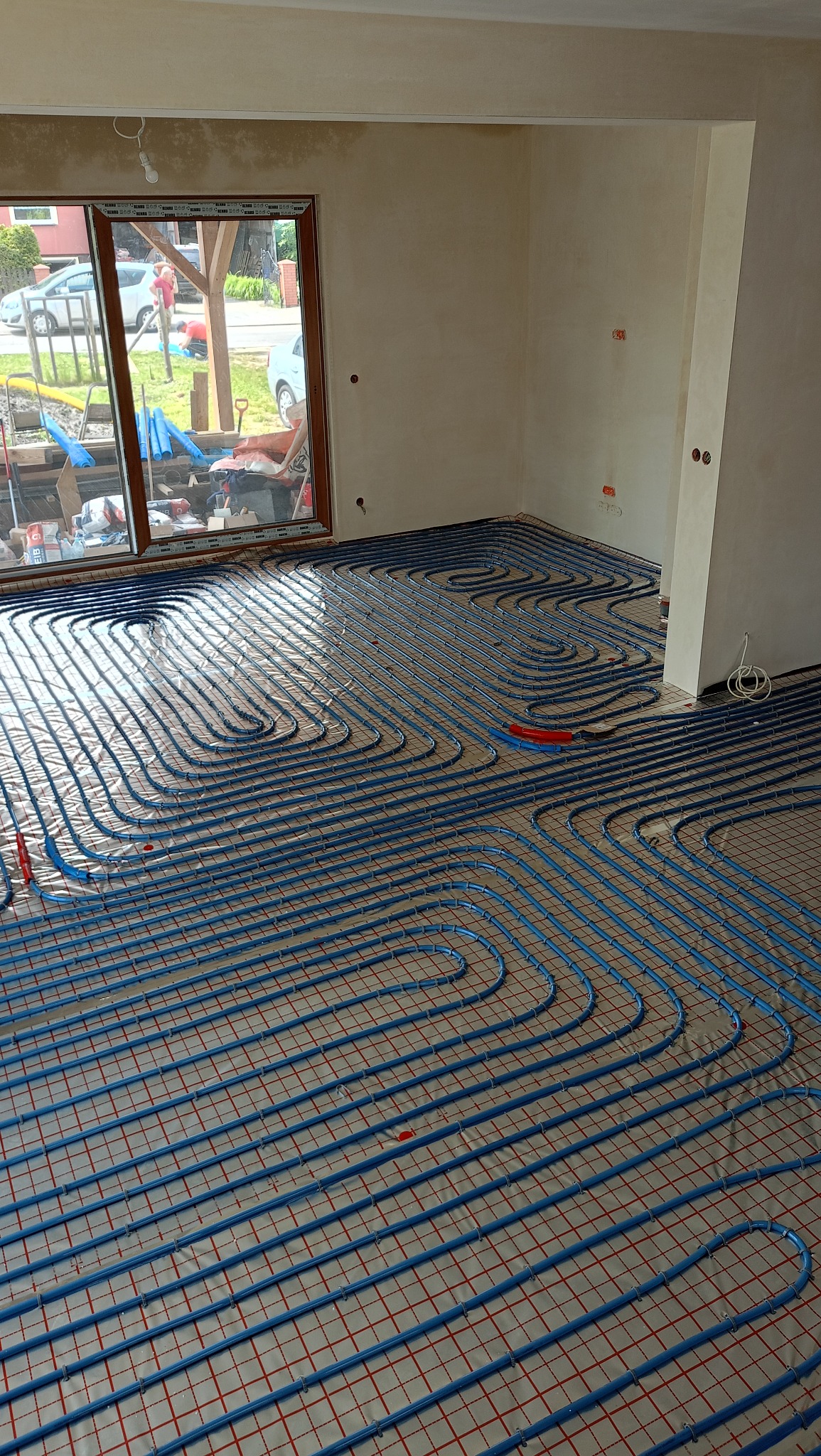 Niebieskie rury ogrzewania podłogowego ułożone na siatce w surowym wnętrzu z oknem. Widoczne detale instalacji i przygotowanie do wylewki.