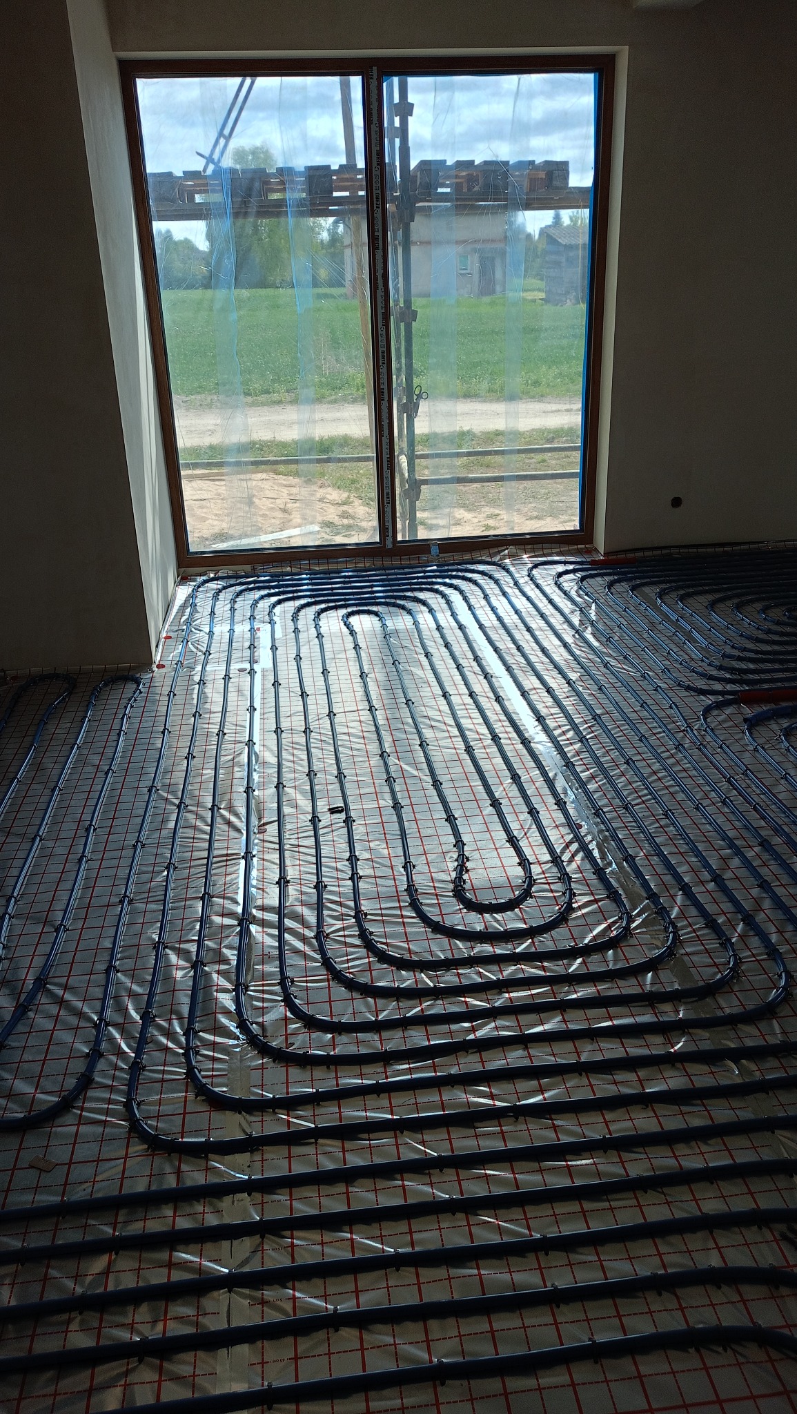 Wnętrze z ułożonym systemem ogrzewania podłogowego na folii, widok przez okno na zewnątrz budynku w budowie, fragment instalacji w prawym górnym rogu.