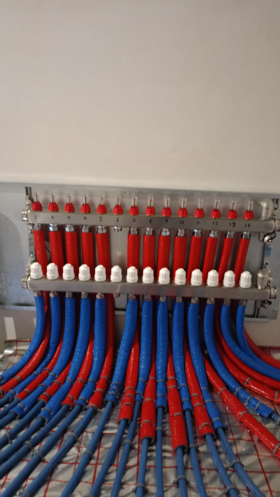 Rozdzielacz ogrzewania podłogowego z rurami w izolacji czerwono-niebieskiej, ułożonymi na macie grzewczej. Widoczne numery obwodów i odpowietrzniki.