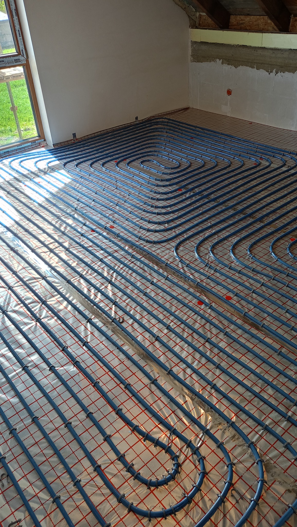 Niebieskie rury ogrzewania podłogowego ułożone spiralnie na folii odblaskowej w surowym wnętrzu z oknem i poddaszem. Widok z góry na etap instalacji.