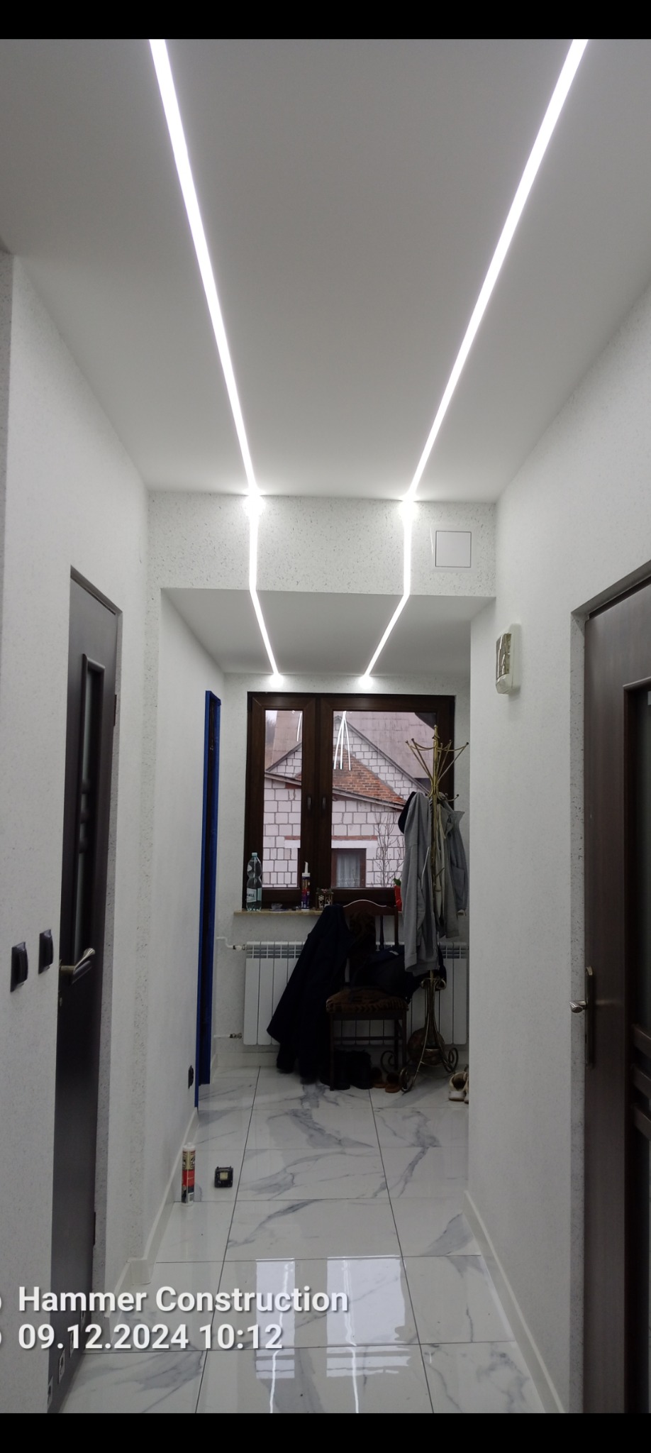 Korytarz po remoncie z białymi ścianami, marmurową podłogą i nowoczesnym oświetleniem LED w suficie. Widoczne drzwi i okno z widokiem na budynek.